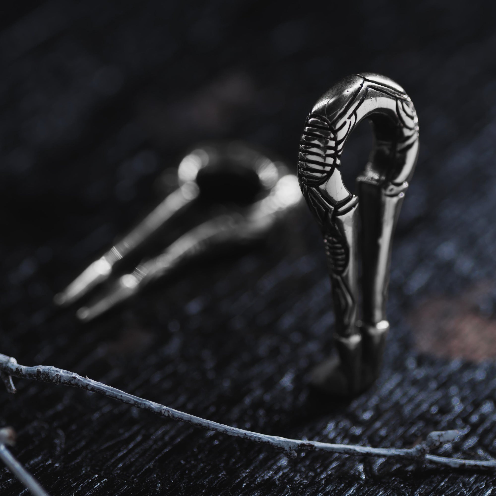 Close-up of the EXRO Biomechanical Cyborg Keyhole Ear Weights in 925 Silver by Kaya Movement, lying on a dark, textured surface. The ear weights exhibit intricate, metallic detailing with a curved frame reminiscent of post-apocalyptic body jewelry. Another set of ear weights is blurred in the background, creating depth. In the foreground, a thin twig is partially visible.